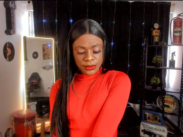 VenusMiller with long black hair, red turtleneck, red lipstick, and eyeliner, looking down, in a room with black curtains and string lights. Boobs not visible.