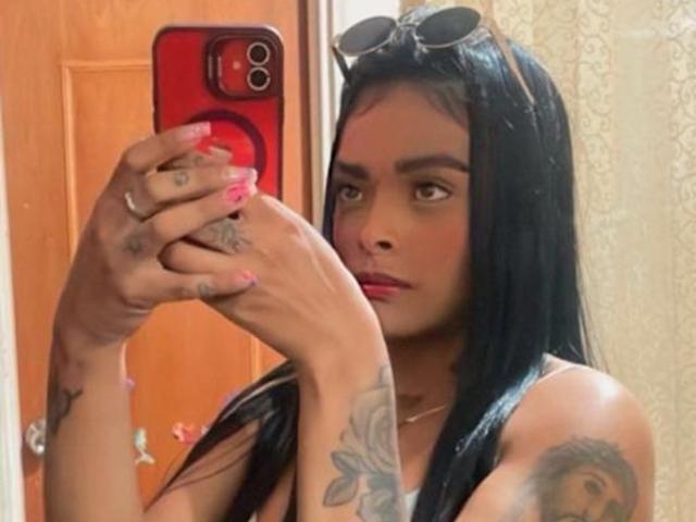 EmmaEbony with long black hair, tattoos on hands and arm, holding red phone, wearing pink lipstick, eyeglasses on head, taking mirror selfie, looking to the side.