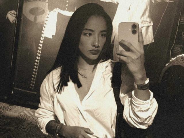 BiancaLeBlanc with long dark hair, wearing a white button-up shirt, taking a selfie with a phone in a dimly lit room, showing her serious face and small tits.