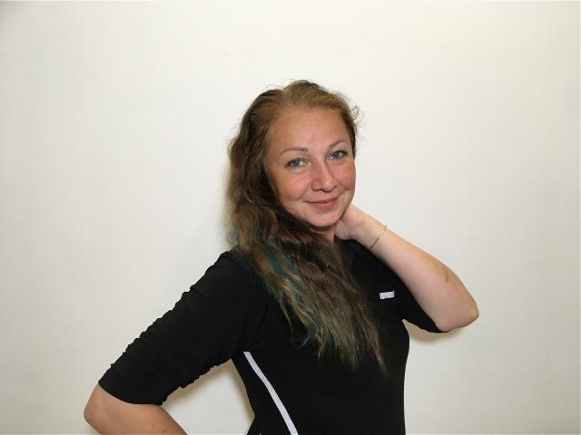 LucyFlirt, with dirty blonde hair, wearing a black top, hand on neck, smiling at camera, has small tits and a cute ass. Plain white background.