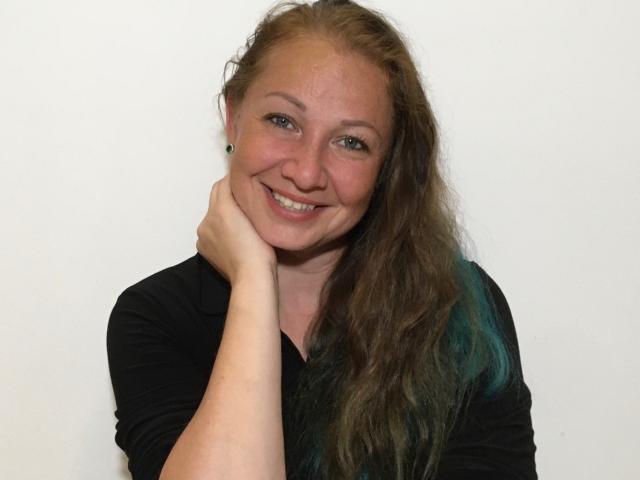 LucyFlirt has long, wavy brown hair with green tips, smiling with her hand on her cheek. She's wearing a black top and has small, cute tits.