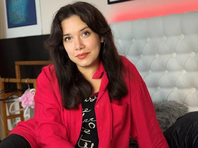 JulietteHayley with dark wavy hair, red shirt, and black patterned top, sitting on a bed, looking at the camera with a slight smile, brown eyes, red lips.