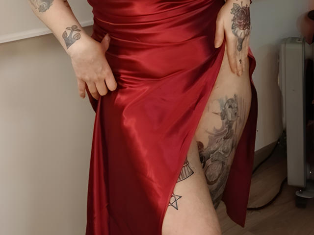 GoddessKrystalViolet in a red satin dress, lifting the skirt to reveal tattoos on her thigh and leg, showing a star and intricate black-and-grey designs.
