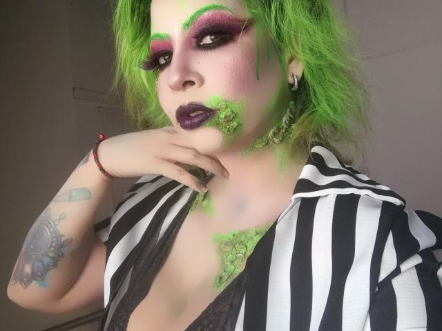 GoddessKrystalViolet with green makeup, purple eyeshadow, green hair, black striped shirt, showing cleavage, hand near mouth, tattoos on left arm, green lipstick, looking at camera.