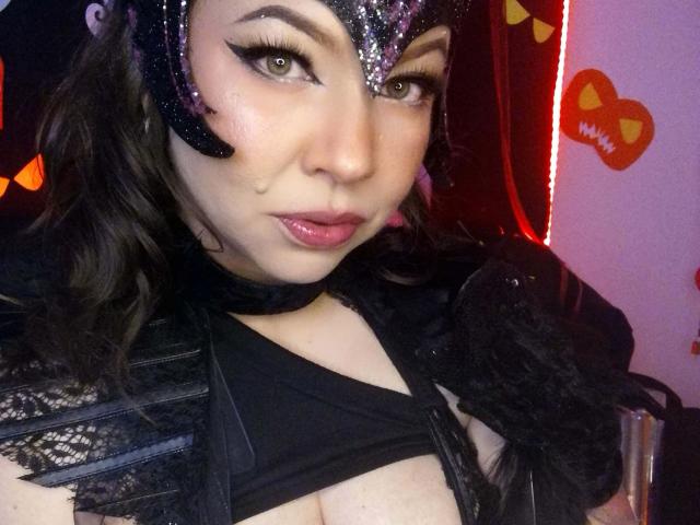 GoddessKrystalViolet in a black, sparkly mask with purple accents, black lace, and a choker, showing her tits in a black top, with pink lips and green eyes.
