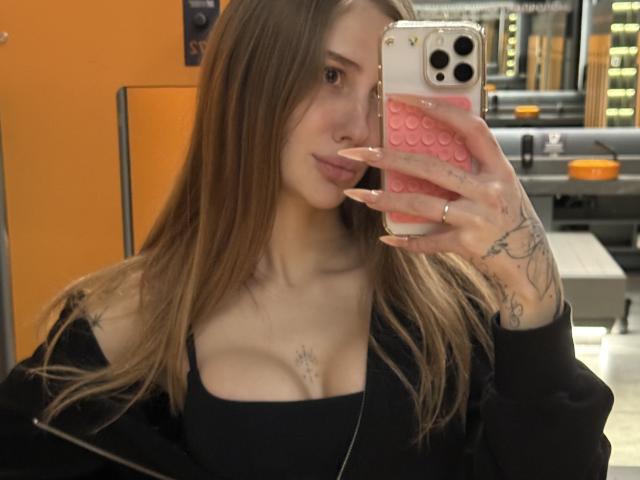 SensualMila with long brown hair, black top, and visible cleavage, taking a mirror selfie with a pink phone case, showing tattoos on her hands and chest.