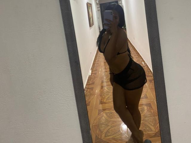 SaraDen in black mesh lingerie, showing her tits and ass, taking a mirror selfie in a hallway, holding a phone, with long dark hair, standing on wooden floor.