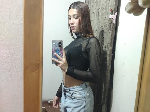 CeelesteBonnett with long brown hair, wearing a sheer black top, showing her small tits and slim midriff, taking a mirror selfie, wearing light blue jeans, holding a phone.