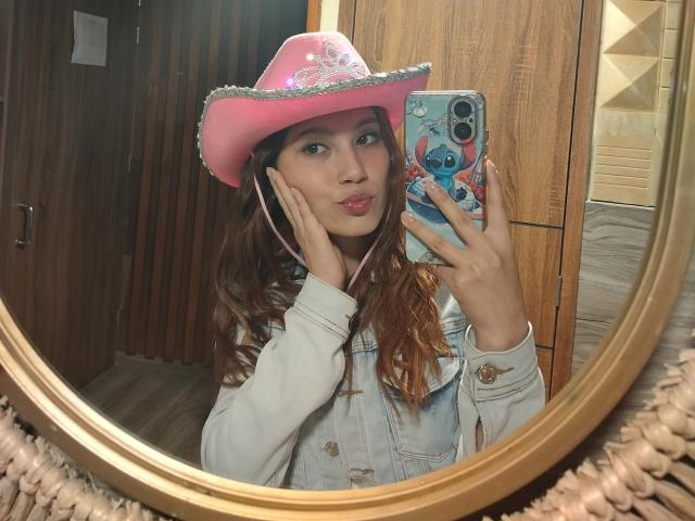 CeelesteBonnett with pink cowboy hat, white sleeves, holding phone with blue Stuffy character, pink earbuds, wavy brown hair, mirror selfie, wooden background, phone case with Stuffy sticker.