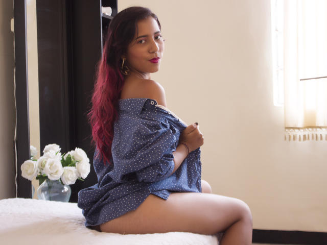 SofiaMarti with red-dyed hair, wearing a blue polka-dot shirt, sitting on a bed, showing her ass, looking over her shoulder, with white roses in the background.