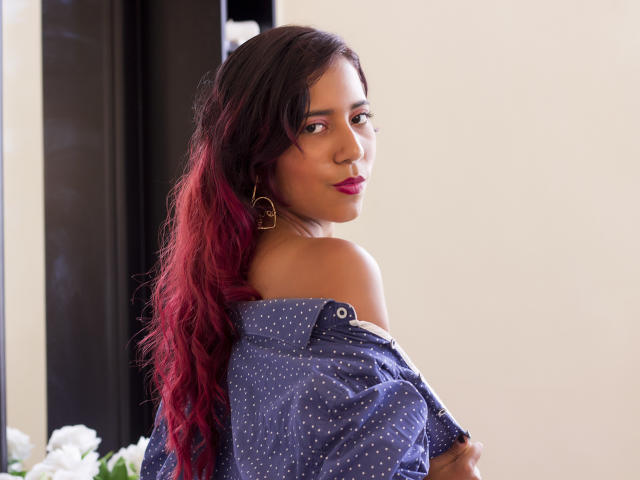 SofiaMarti with long red-to-black ombre hair, off-shoulder blue polka dot shirt, gold heart earrings, looking back with pink lipstick, showing bare shoulder and upper chest.
