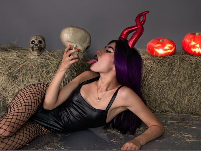 JuliaJobs with long purple hair, black shiny dress, and red devil horns, lies on gray floor, licking a skull. She wears black fishnet stockings, has orange nail polish, and a gold necklace. Behind her, hay bale with skull and three glowing red pumpkins. The composition is dark and Halloween-themed, with a focus on JuliaJobs' provocative pose and the eerie, seasonal props. Textured hay contrasts with her shiny dress. 

 