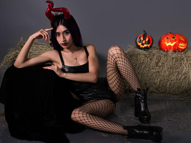 JuliaJobs mit schwarzen Hörnern, lila Haaren, einem schwarzen Lederkleid, Fischernetzstrümpfen und schwarzen Stiefeln, sitzt auf Heu mit zwei leuchtenden Halloween-Kürbissen. Brüste, Hintern, Beine und Vulva sind sichtbar..