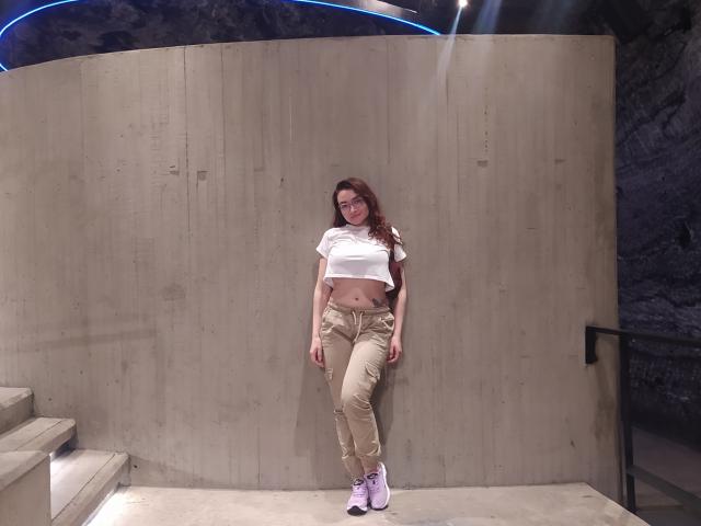 Drikka stands against a concrete wall, wearing a white crop top, beige pants, and pink sneakers. Her long brown hair is loose, and she has a small belly tattoo.