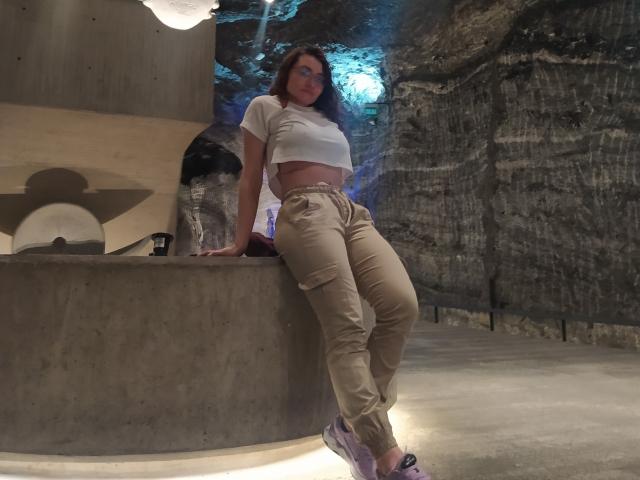 Drikka, con un seno di media grandezza, indossa una canotta bianca, pantaloni beige e scarpe da ginnastica viola, appoggiata a una superficie di cemento in una stanza a forma di grotta, scarsamente illuminata..