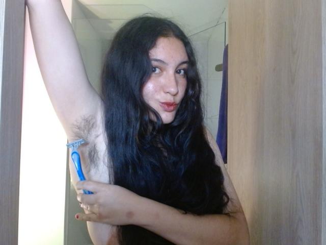MargaritaDudole with long black hair, pouty lips, holding a blue razor in her right hand, shaving her hairy left armpit. She has a light skin tone and a subtle smile.