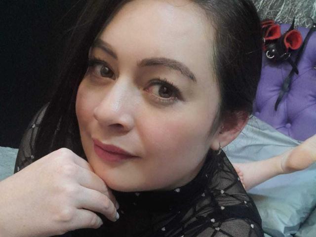 KristelRouse's face with dark brown hair, light skin, and subtle makeup, wearing a black sheer top, resting her chin on her hand, with a slight smile. Her pussy is off-frame.