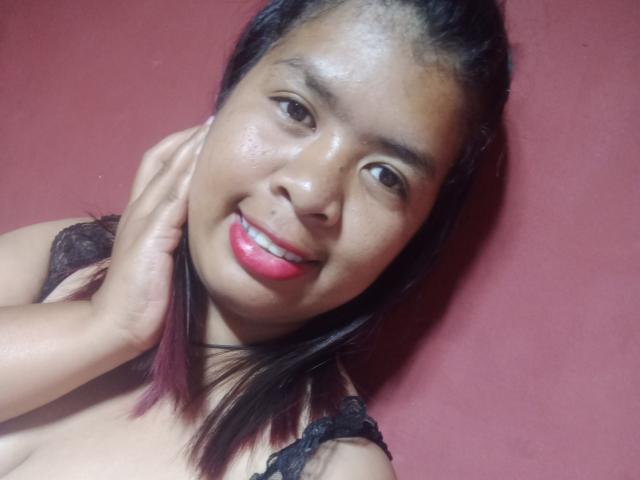 MariahSquirting69 has dark brown skin, black lace bra, pink lipstick, smiling, hand on neck, black hair with purple tips, and visible small breasts against a red background.