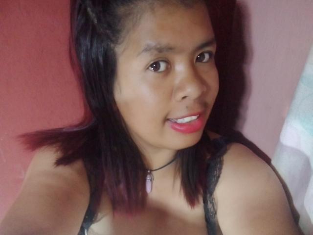 MariahSquirting69 with dark brown eyes, red lipstick, black lace bra, and a pink stone necklace, smiling at the camera, showing her tits, against a pink wall.