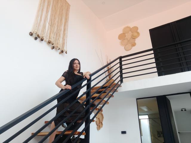 MargaritaDudole, with black dress and long black hair, stands on a black metal and wooden staircase, hand on railing, looking down, with black and white walls behind.