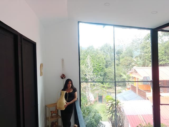 MargaritaDudole with black hair, black dress, holding yellow bag, standing near window, white wall, dark door, wooden shelf, and small basket in modern room, looking outside.