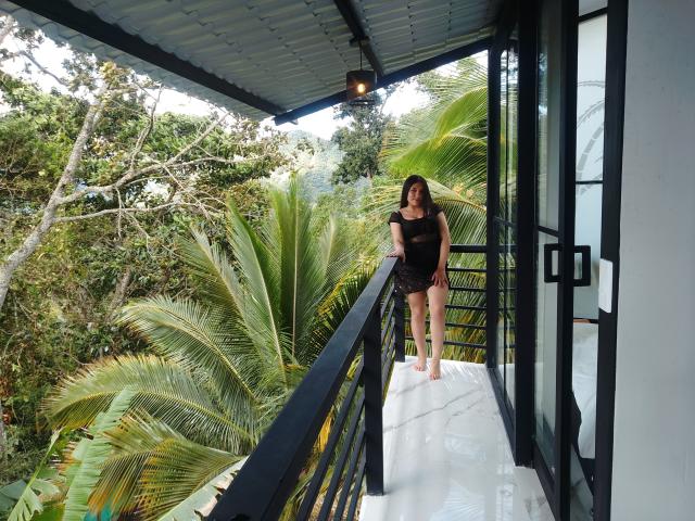 MargaritaDudole stands on a white marble balcony with black railings, wearing a black sheer top and black shorts, surrounded by lush green palm trees and a metal roof.