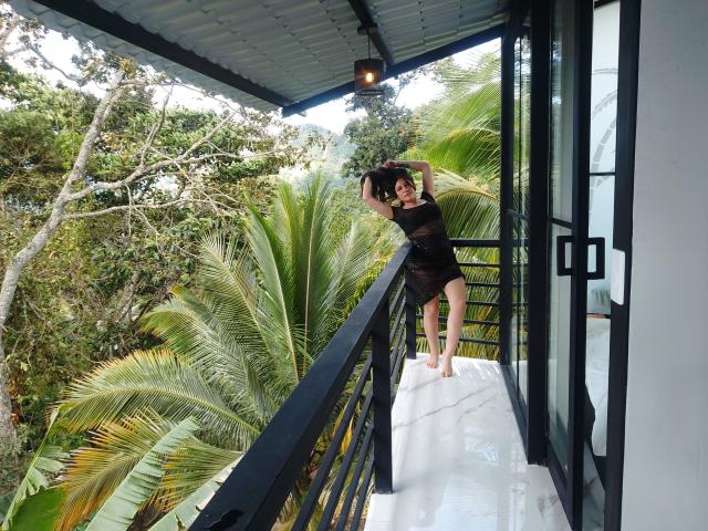 MargaritaDudole with curly black hair, wearing a black sheer dress, standing on a balcony, arms above head, black railing, glass door, palm trees, and overcast sky in background.