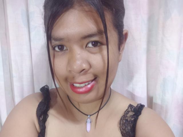 MariahSquirting69 tiene ojos marrones, lápiz labial rojo, un sujetador de encaje negro y un collar de piedra morada, y está sonriendo a la cámara. Su cabello negro está recogido en un moño suelto..