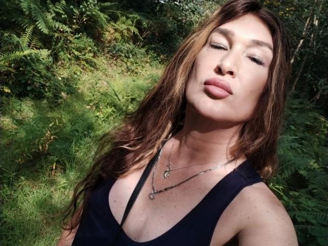 Tsnovaleex, avec ses lèvres pulpeuses, ses cheveux bruns ondulés et son débardeur noir, porte un collier et se trouve dans une forêt verte. Elle a un joli visage et de petites seins..