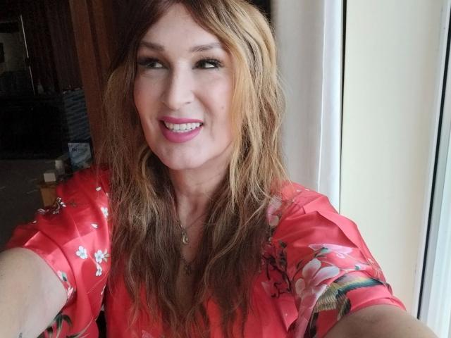 Tsnovaleex, avec ses cheveux bruns ondulés, sa robe fleurie rose et son rouge à lèvres rose, sourit à l'appareil photo, montrant ses dents, et tend son bras pour un selfie. Ses seins sont légèrement visibles sous la robe..