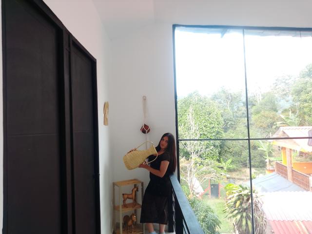 MargaritaDudole with long dark hair, black dress, holding wicker bag, standing near large window, white wall, black door, wooden shelf, green tree-filled view outside, bright daylight.
