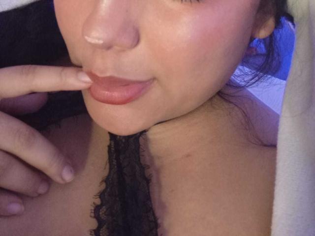 KonyBella's face, showing pink lips, a slightly shiny nose, and a black lace choker. She's touching her lip with her finger, and her hand is near her chest.