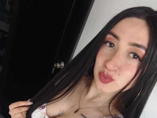 JuliethCruz with long black hair, pink eyeshadow, red lipstick, pouty lips, small tattoos on chest, wearing off-shoulder top, holding hair, large hoop earrings, white wall in background.