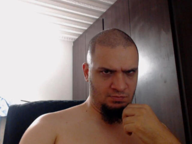 Shirtless Sigfridd with a bald head and a beard, looking angry, holding his chin. His eyes are squinted, and he's making a slight frown. Dark wooden cabinet behind.