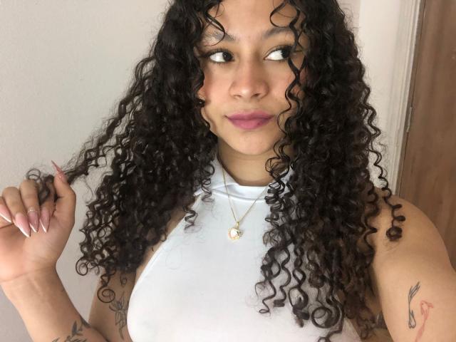 SofiaPassionne has curly black hair, looking to the side with a slight smile. She wears a white sleeveless top and a gold necklace with a small pendant. Her right hand is raised, touching her hair. She has light brown skin, pink lipstick, and visible tattoos on her right arm. Her nails are long and painted pink. The image focuses on her upper body.