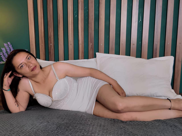MiahDurand with medium tits in a white lace dress, lying on a bed with gray blanket, wooden slats behind, white pillows, brown hair, green eyes, and a relaxed expression.