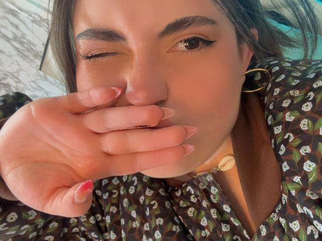 VicluX with one eye closed, hand covering mouth, wearing a brown floral shirt, gold hoop earrings, and a shell necklace, has long nails and a cute winky face.