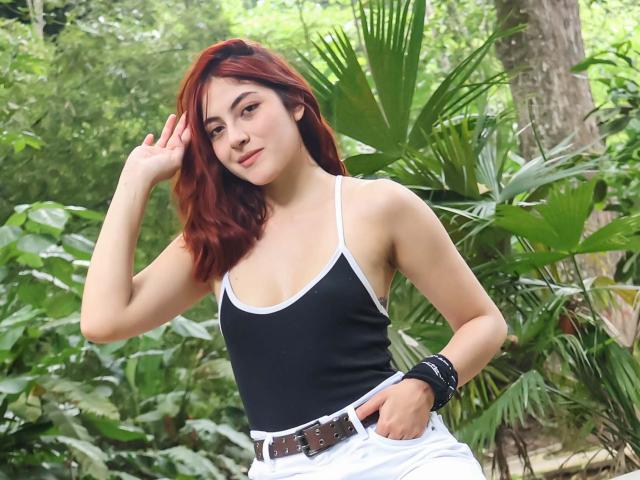 SpencerLe met rood haar, gekleed in een zwart tanktop en witte broek, met een hand in de zak, een zwarte armband, staand in een groene jungle, kijkend naar de camera, met een lichte glimlach..