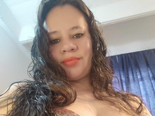 A close-up selfie of CharllotI with wet, wavy brown hair cascading over her shoulders. She has light brown skin, brown eyes, and is wearing orange lipstick. Her expression is neutral with a slight smile. She's wearing a red-patterned top. The background has a blue curtain and white ceiling. The lighting is soft, highlighting her face. The image has a casual, everyday feel. The composition is centered, focusing on CharllotI's face and upper torso.