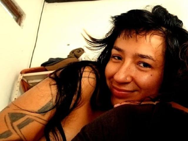 CassandraMorelo with black hair, nose ring, tattooed arm, smiling at camera, holding brown pillow, in dimly lit room.