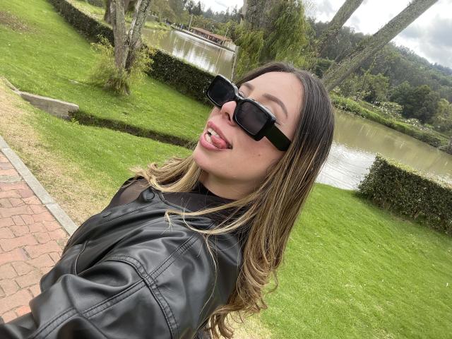 JennyLawry with tongue out, black sunglasses, and a black leather jacket, showing off her long brown hair and green grass park in the background. Her ass is not visible.