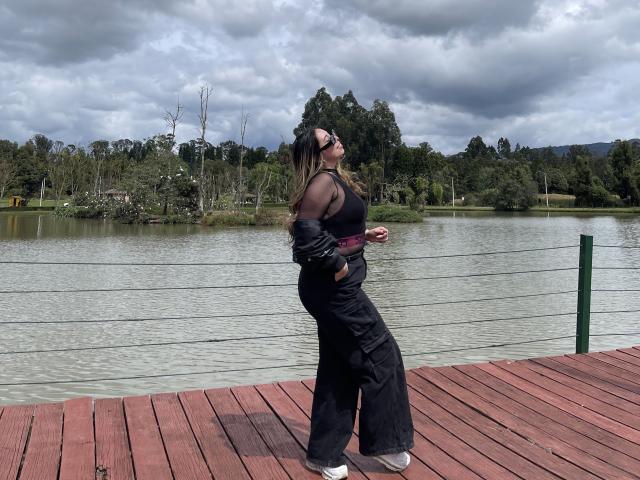 JennyLawry stands on a wooden dock, wearing a black tank top, black pants, and sunglasses, with her hands in her pockets, looking to the side, under a cloudy sky.