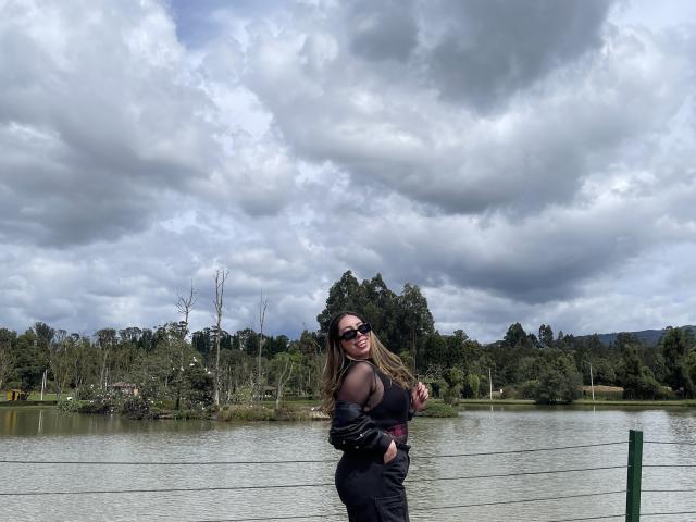 JennyLawry, con grandes senos, trasero curvilíneo, cabello largo y castaño, gafas de sol negras, top negro transparente, chaqueta de cuero negro, de pie junto a un lago, cielo nublado, árboles verdes en el fondo..
