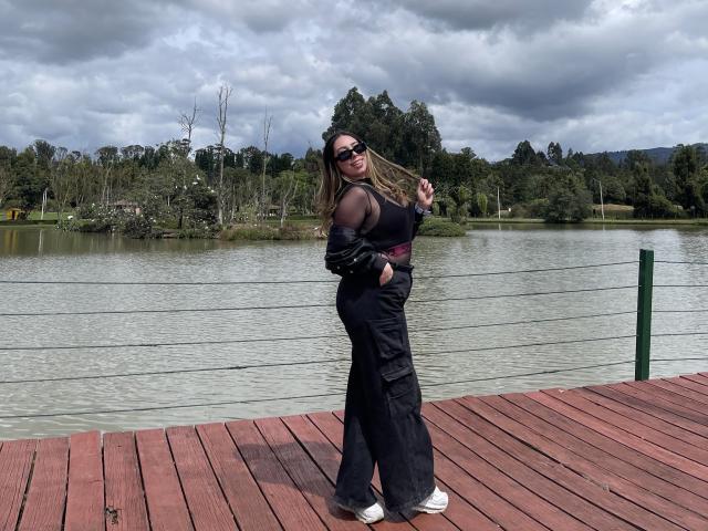 JennyLawry with medium tits, wearing black cargo pants, black top, sunglasses, and white sneakers, standing on red wooden dock, holding jacket, near lake with green trees in background.