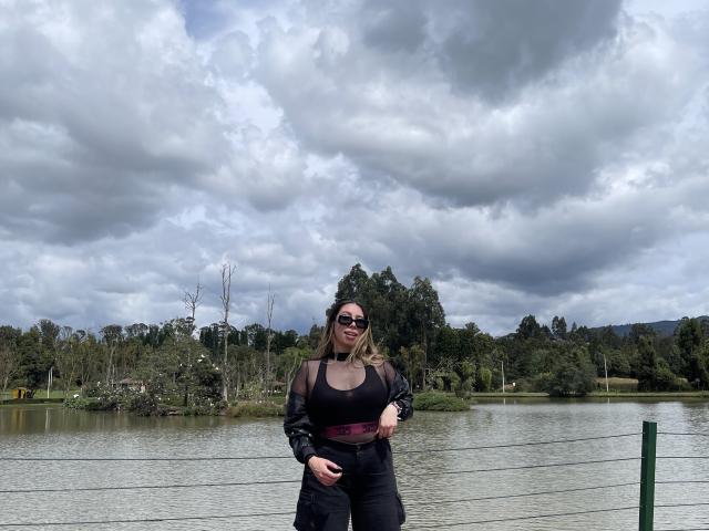 JennyLawry with big boobs, wearing black sheer top, black pants, maroon belt, sunglasses, standing by a wire fence, next to a lake with cloudy sky.