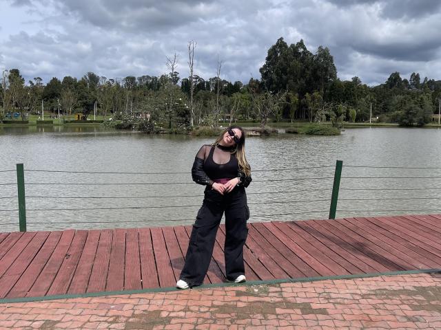 JennyLawry stands on a wooden dock, wearing black clothes, glasses, and white shoes, with hands on her ass, looking up, cloudy sky, green fence, and lake in background.