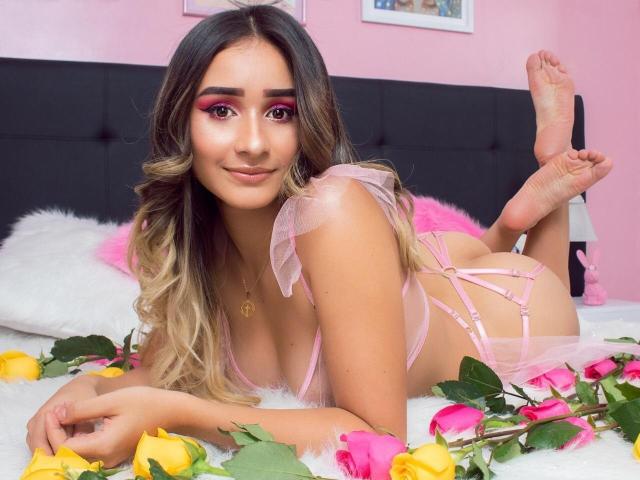 LauraKlark with long, wavy blonde hair, pink eyeshadow, and pink lingerie, lying on a bed, legs up, surrounded by yellow and pink roses, showing her ass.
