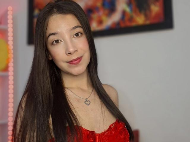 ManuMejia with long black hair, red lipstick, and a red lace top, wearing a heart necklace, smiling slightly. Boobs visible under lace top. Soft, light skin.