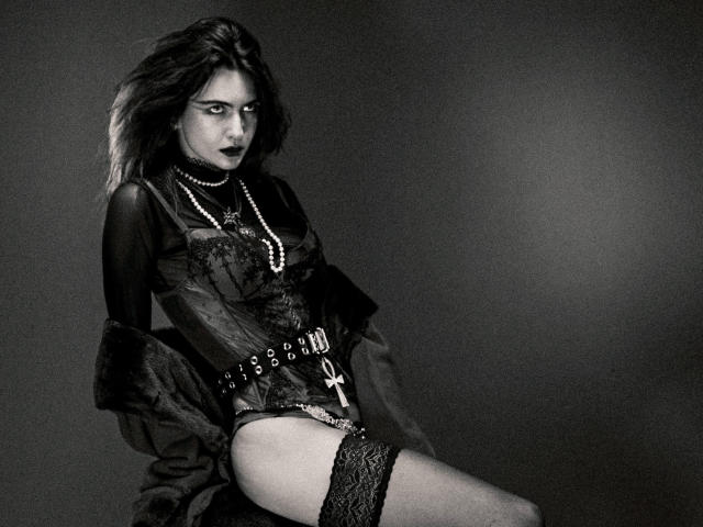 CosmoAngy with dark hair, wearing a black lace-up corset, pearl necklace, black lipstick, and lace thigh-highs, sitting and looking seductively at the camera. Black-and-white.