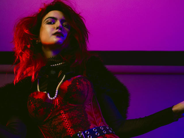 CosmoAngy, con cabello rojo, vistiendo un corpiño de encaje negro, estampado floral rojo, collar de perlas, chaqueta de piel negra y un cinturón con anillos de metal, iluminada por luces moradas y rojas..